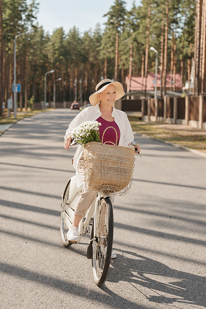 Frau mit Sonnenhut fährt an einem sonnigen Tag mit dem Rad, im Korb ein Strauß weißer Blumen